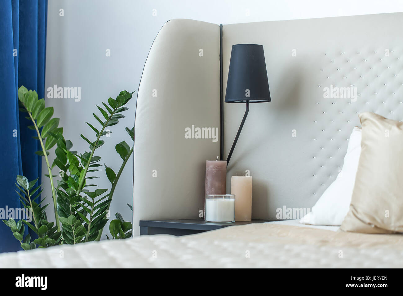 Bett mit Kissen auf die Lichtwand Hintergrund. Auf der linken Seite gibt es eine dunkle Nighstand mit Kerzen und einer Lampe, Grünpflanze, blauen Vorhang. Closeup. Indo Stockfoto