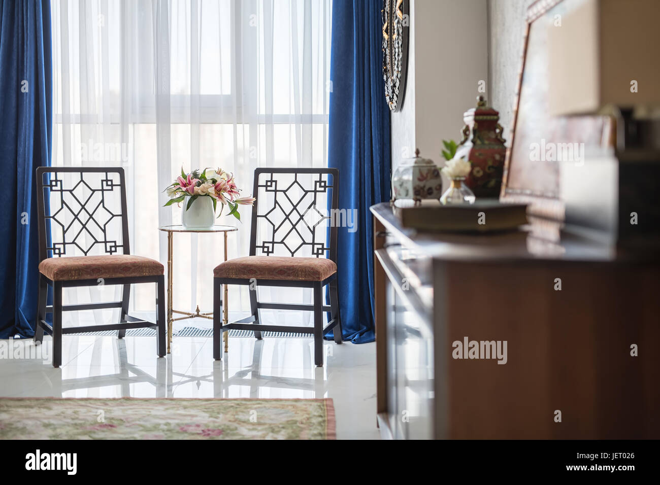 Interieur in einem modernen Stil mit hellen Wänden und einem gefliesten Boden mit einem Teppich. Es gibt zwei stilvolle Stühle, runder Tisch, Blumen in Vasen, ausgefallene Spiegel Stockfoto