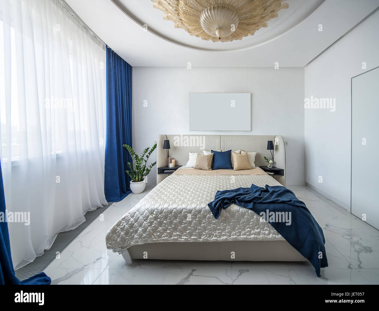 Moderne Schlafzimmer mit einem großen Phantasie goldene Kronleuchter an der Decke. Gibt es eine Beige Bett mit bunten Kissen und ein blaues Karomuster, Regale mit Kerze Stockfoto
