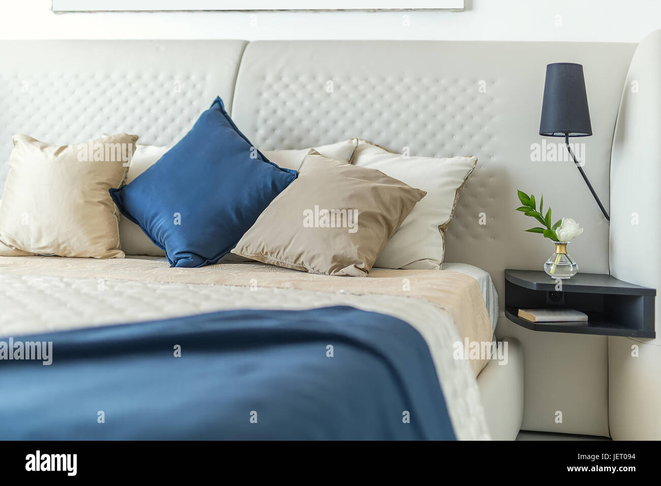 Breites Bett mit bunten Kissen und Badetücher auf die weiße Wand Hintergrund. Auf der rechten Seite ist ein schwarze Regal mit einer Blume in einer Glasvase und eine Stockfoto