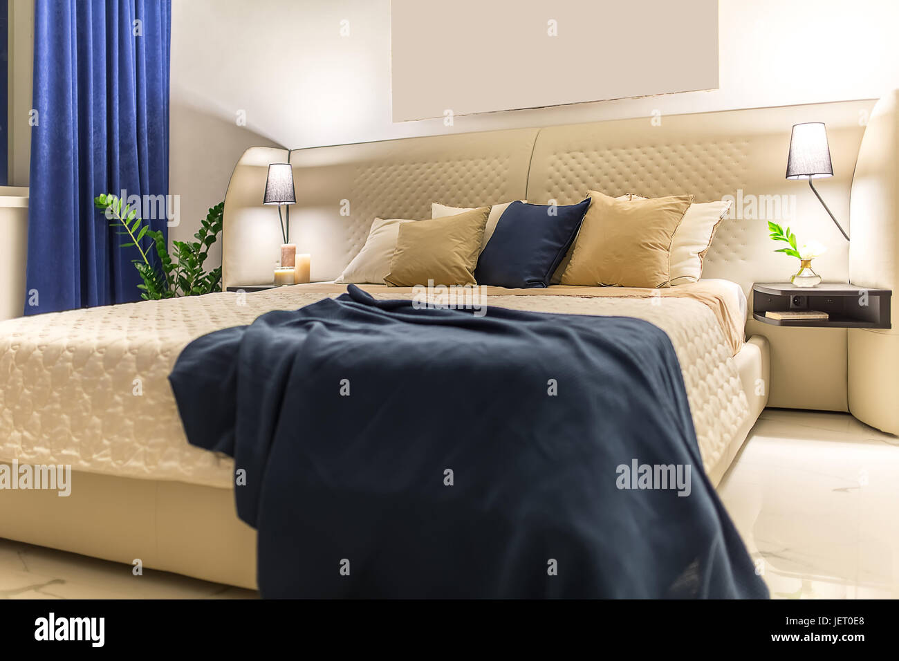 Helle moderne Schlafzimmer mit weißen Wänden und Licht gefliesten Boden. Es ist ein Beige Bett mit bunten Kissen und ein blaues Karomuster, Regale mit Kerzen Stockfoto