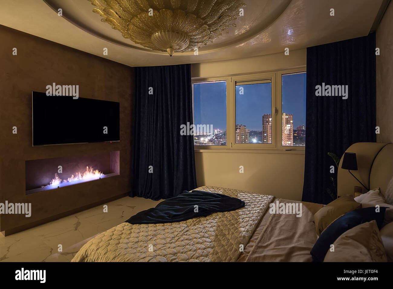 Schlafzimmer im modernen Stil mit einem großen Phantasie goldene Kronleuchter an der Decke. Es ist ein Bett mit Kissen und eine Tagesdecke, Kamin, TV auf dem Stockfoto