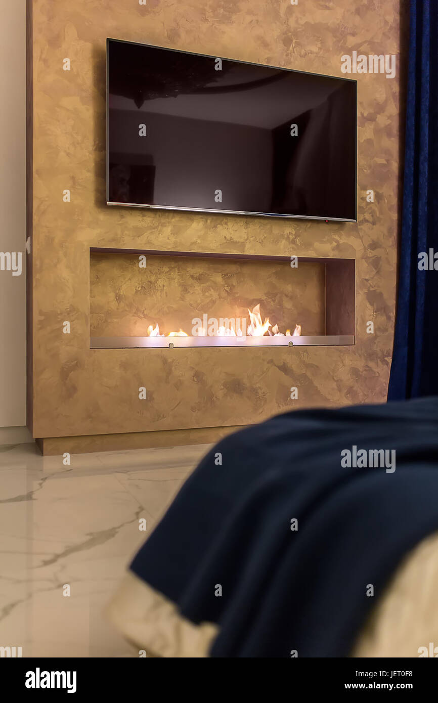 Strukturierte Bronze Wand mit einem Kamin mit einem brennenden Feuer und einen großen Fernseher. Es ist ein Bett mit einer blauen Tagesdecke vor der Wand. Auf der Etage gibt es eine Stockfoto
