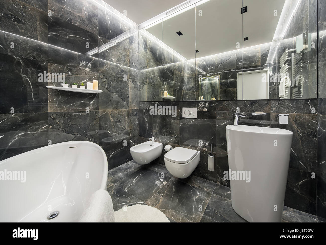 Modernes Badezimmer mit strukturierten dunklen Fliesen und leuchtenden Lampen. Es ist ein weißes Bad, großem Spiegel, WC und Bidet, großer Spüle mit einem Chrom-Armatur, Stockfoto