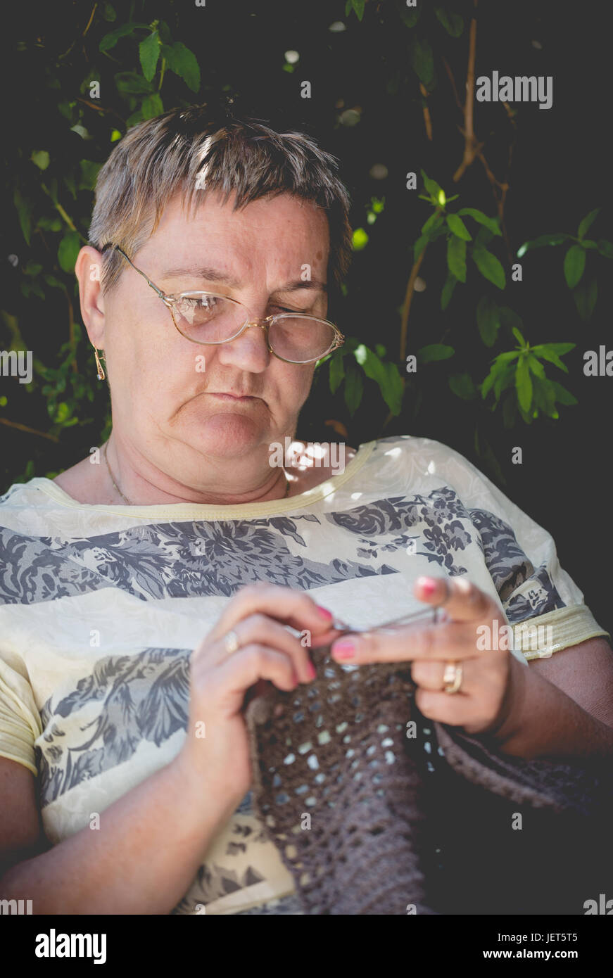 Ältere Frau, kurze Haare, häkeln eine braune Decke entspannend im Garten. Vertikale Zusammensetzung Schwerpunkt konzentrierte Gesicht Stockfoto