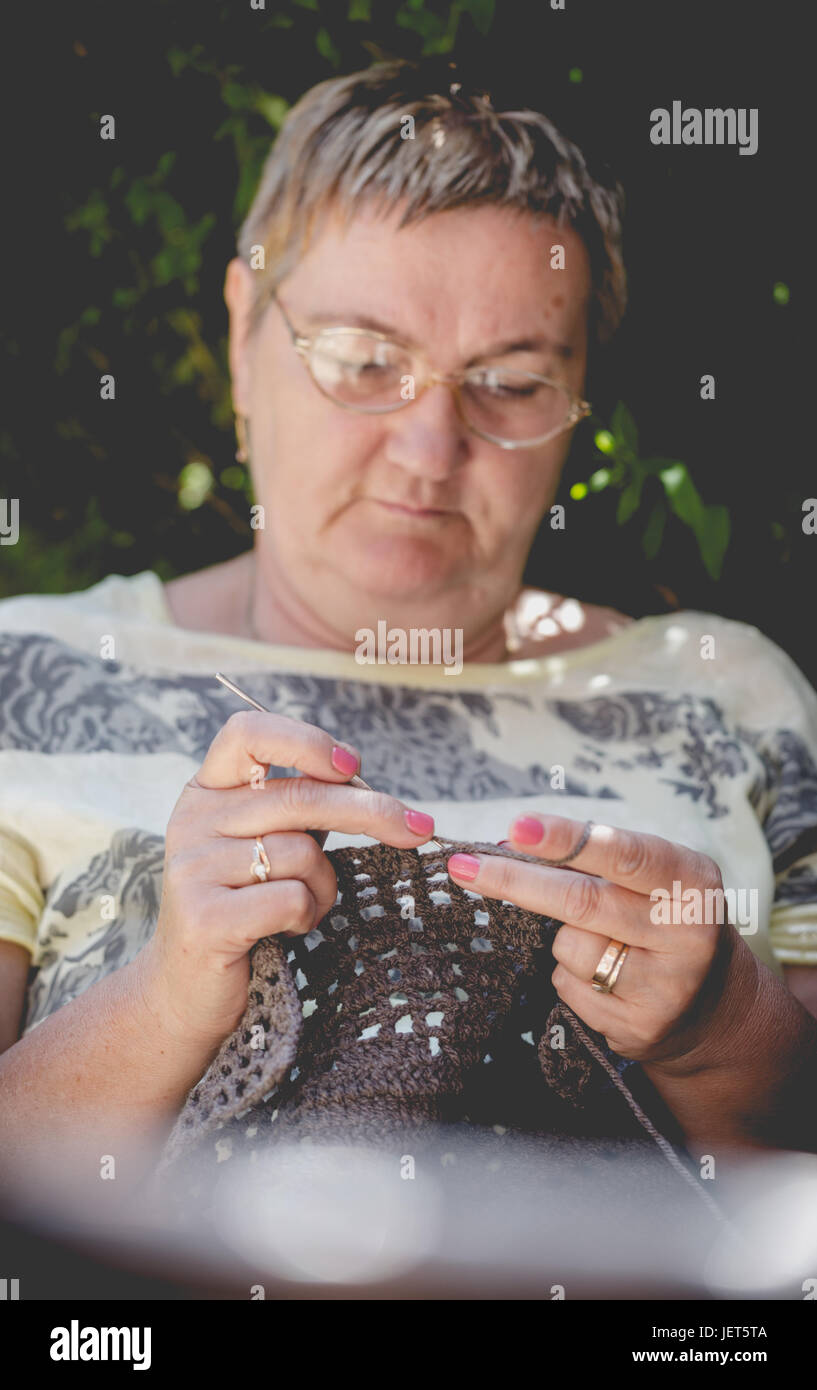 Ältere Frau, kurze Haare, häkeln eine braune Decke entspannend im Garten. Vertikale Zusammensetzung Schwerpunkt arbeitende Hände Stockfoto