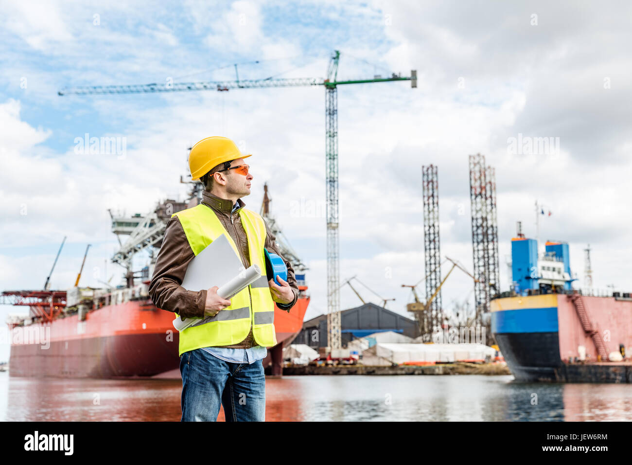 Schiffbau-Ingenieur steht am Hafen in einem Hafen. Gelbe Weste, Helm und Schutzbrille tragen. Schiffbauindustrie. Stockfoto