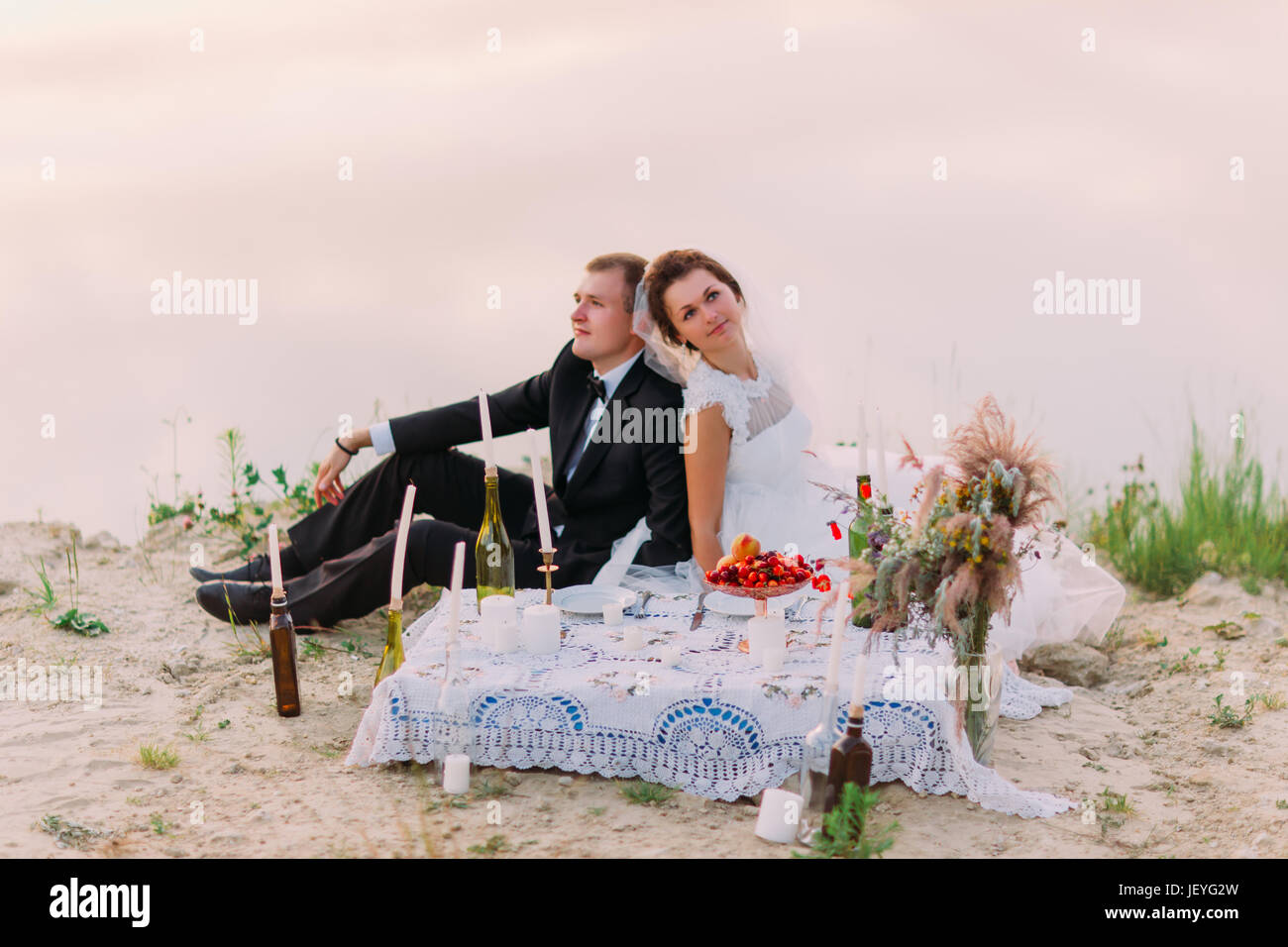 Das romantische horizontale Porträt von dem Brautpaar Sirring Rücken an Rücken während der Hochzeit Picknick auf der Küste Hochzeit Picknick an der Küste Stockfoto