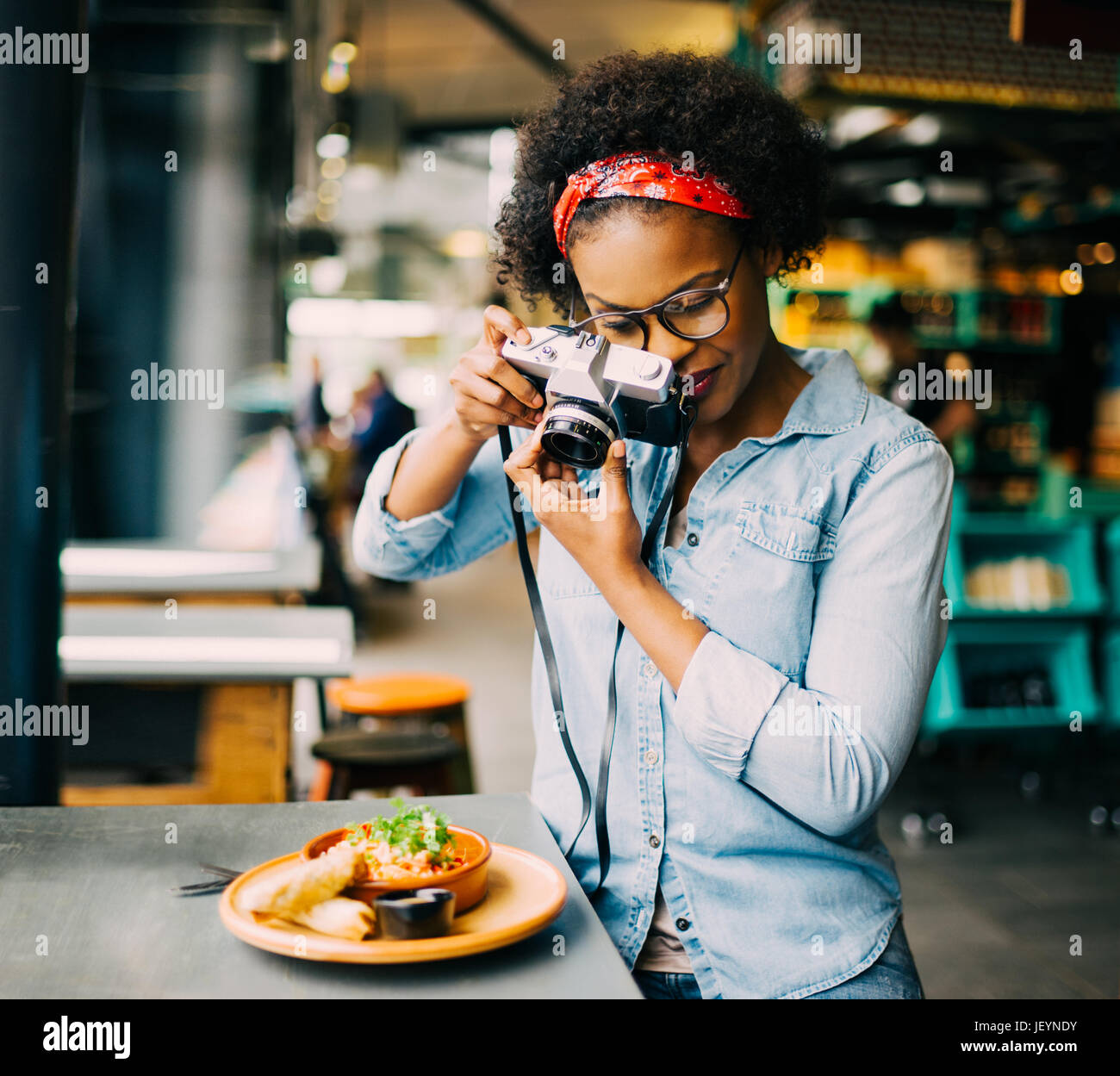 Junge afrikanische Frau allein an einem Schalter in einem Bistro, die Fotos von ihr Essen mit einem Vintage slr-Kamera Stockfoto