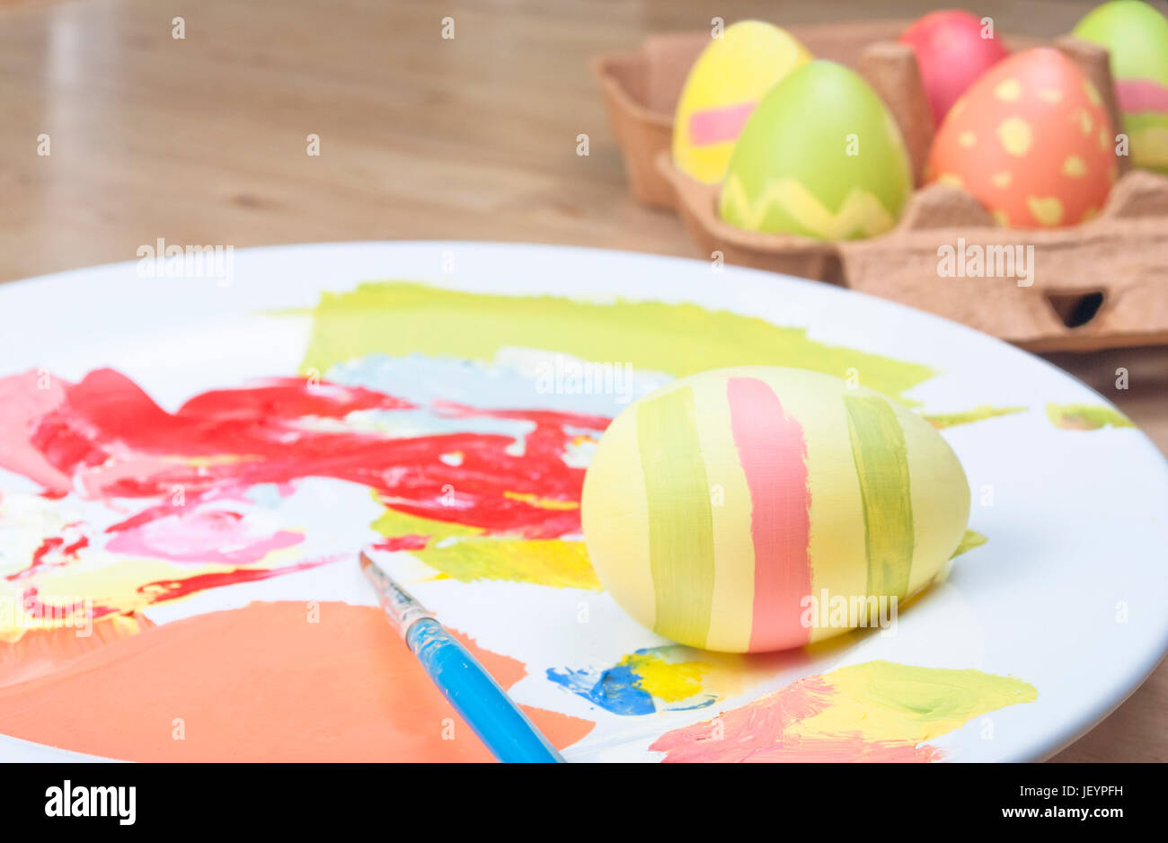 Eine gemütliche Osterei-Malerei-Szene auf einem Holztisch.  Fertige Eiern in einem Karton, Ei-in-Progress, Farben und Pinsel auf einer China-Teller-Palette in Stockfoto