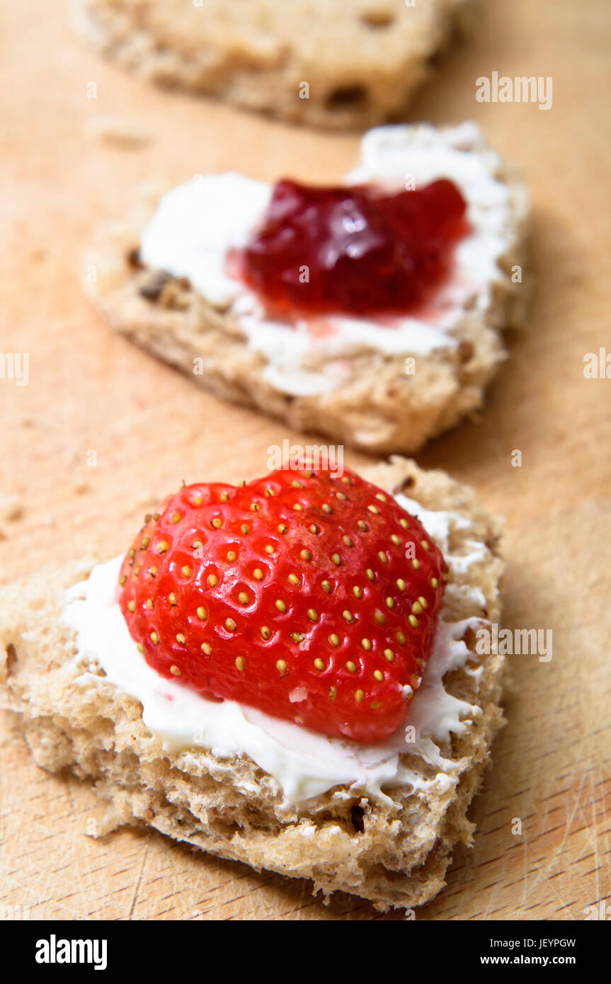 Eine vertikale Reihe von Herzen geformt Getreidespeicher Brot Snacks auf ein Schneidbrett aus Holz.  Belag Frischkäse, Erdbeeren und Marmelade. Stockfoto