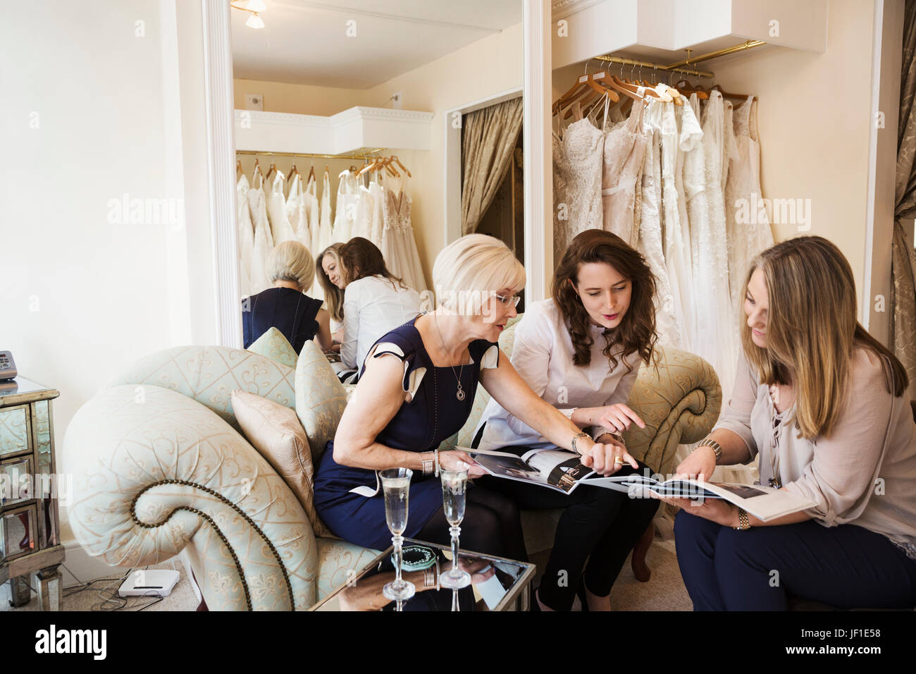 Drei Frauen in einer Hochzeit Kleid Shop, eine Braut zu sein und zwei Retail Berater Blick durch Broschüren und Zeitschriften. Zwei Gläser Champagner auf der Stockfoto