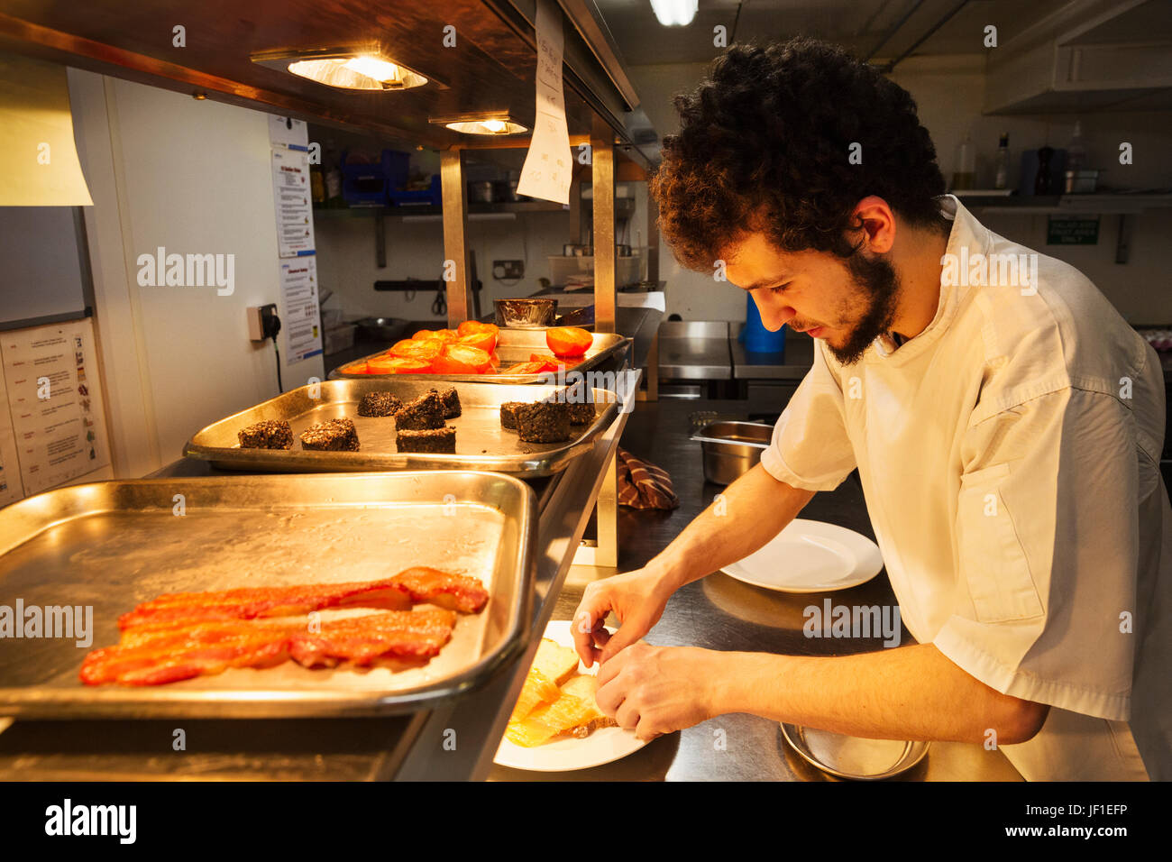 Koch in einer Restaurantküche bei einem Zähler Beschichtung Essen, Tabletts mit Speck, Blutwurst und gegrillten Tomaten stehen. Stockfoto