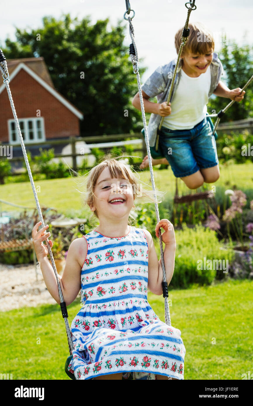 Lächelnde junge in Hemd und Jeans Shorts und Mädchen in einem Sommerkleid auf Schaukel im Garten. Stockfoto