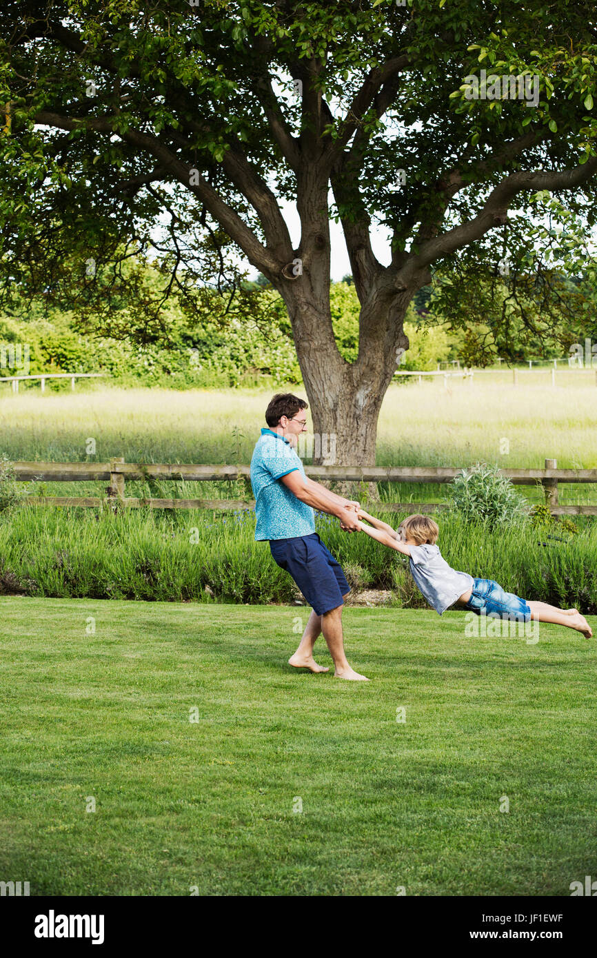 Mann, stehend auf einer Wiese in einem Garten, ein Junge von seinen Händen hält, Spinnen und wirbeln ihn herum. Stockfoto