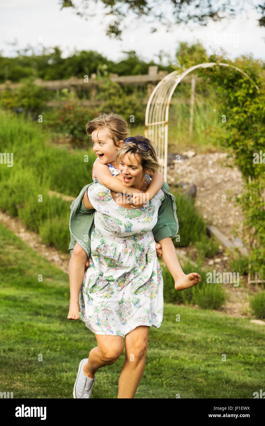 Frau in einem Sommerkleid quer über eine Wiese geben einem Kind eine Huckepack. Stockfoto