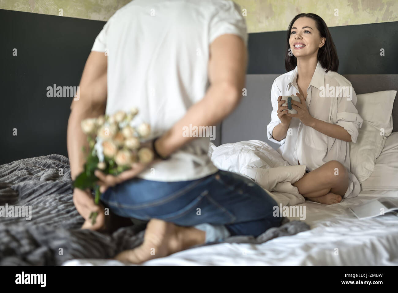 Mann in Blue Jeans und weißes T-shirt hält einen Strauß Rosen hinter seinem Rücken. Er sitzt auf dem Bett gegenüber seiner lächelnden Freundin in ein weißes Hemd mit Stockfoto