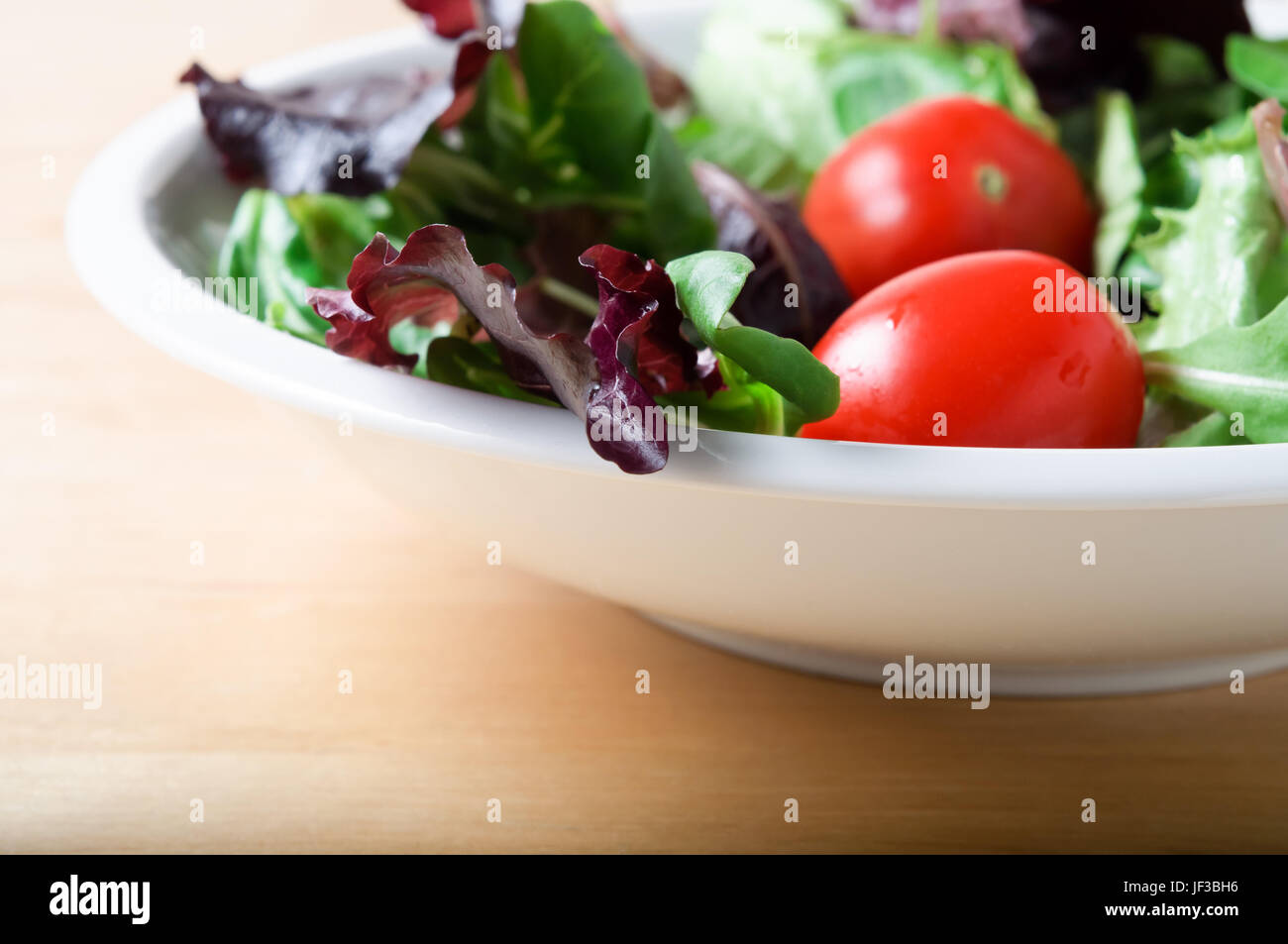 Eine weiße Schüssel mit Salat, einschließlich roter Kopfsalat, Rucola und Tomaten auf einem Leuchttisch aus Holz.  Querformat (horizontale). Stockfoto