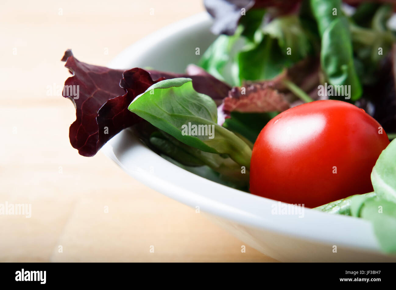 Nahaufnahme (Makro) roter Kopfsalat, Rucola und Kirschtomaten Salat in einer weißen China Schüssel auf einem Leuchttisch aus Holz.  Querformat (horizontale). Stockfoto