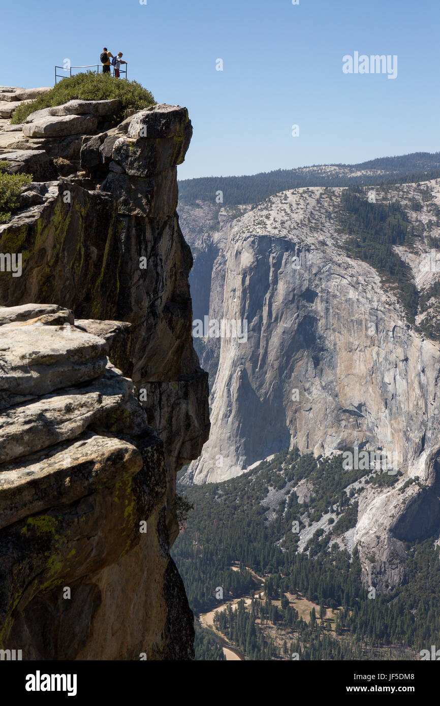 Mit El Capitan in der Ferne am Taft Point wandern trail Peak, Wanderer Stand auf einem Geländer und schauen über den Rand zum Yosemite Tal unten. Stockfoto