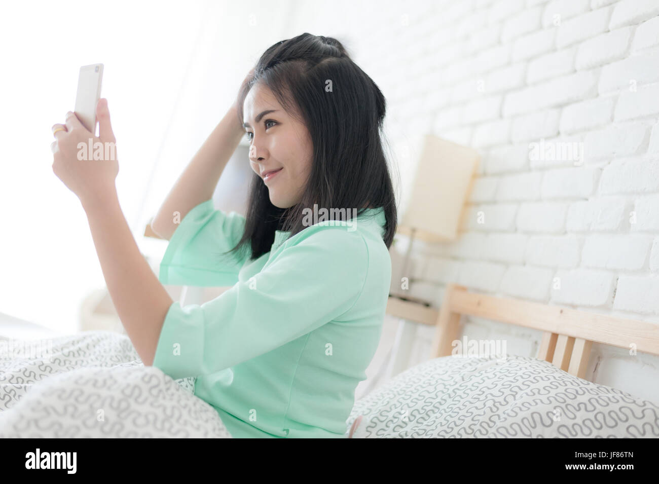 Junge attraktive Asiatin tragen grüne lässige Tücher Selfie Aufnahme beim Sitzen auf dem Bett. Faule Tätigkeit am Sonntagmorgen. Stockfoto