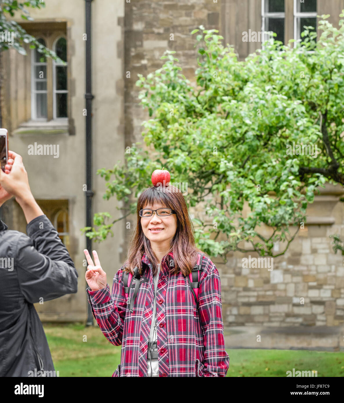 Weibliche chinesische Touristen posiert mit einem Apfel auf dem Kopf vor einem Apfelbaum am Trinity College in Cambridge, wo Isaac Newton lebte und lehrte. Stockfoto