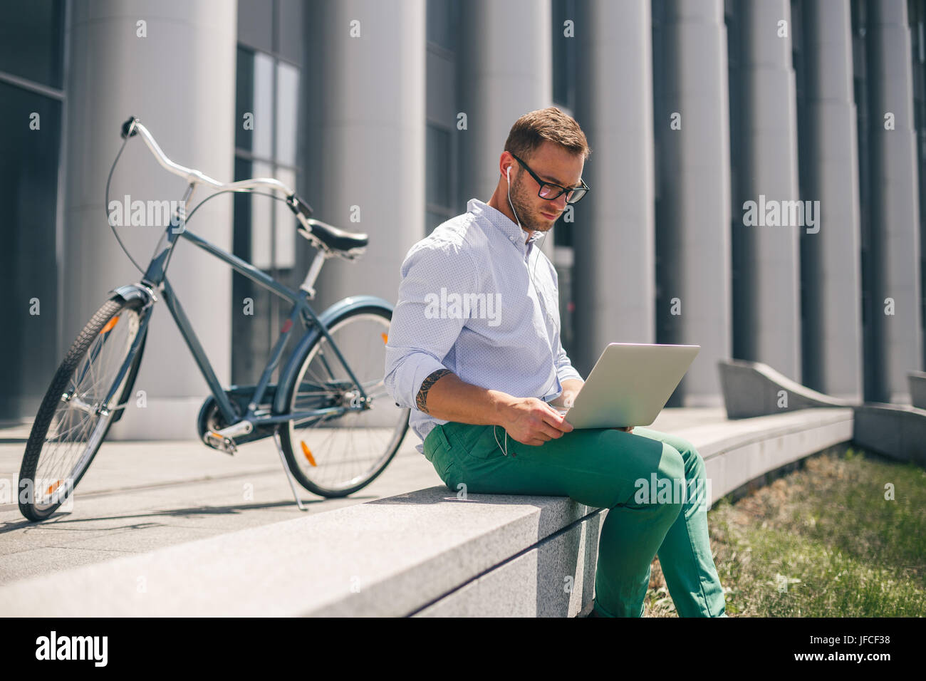 Junger Mann im Kopfhörer am Laptop an Stadtstraße arbeiten. Lifestyle, Verkehr und Menschen Konzept. Im Hintergrund ist ein Fahrrad. Stockfoto