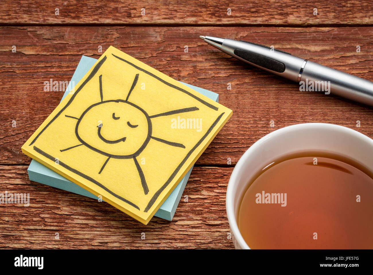fröhlich lachende Sonne - Skizze in schwarzer Tinte auf einem gelben Zettel gegen rustikales Holz mit Tee Stockfoto