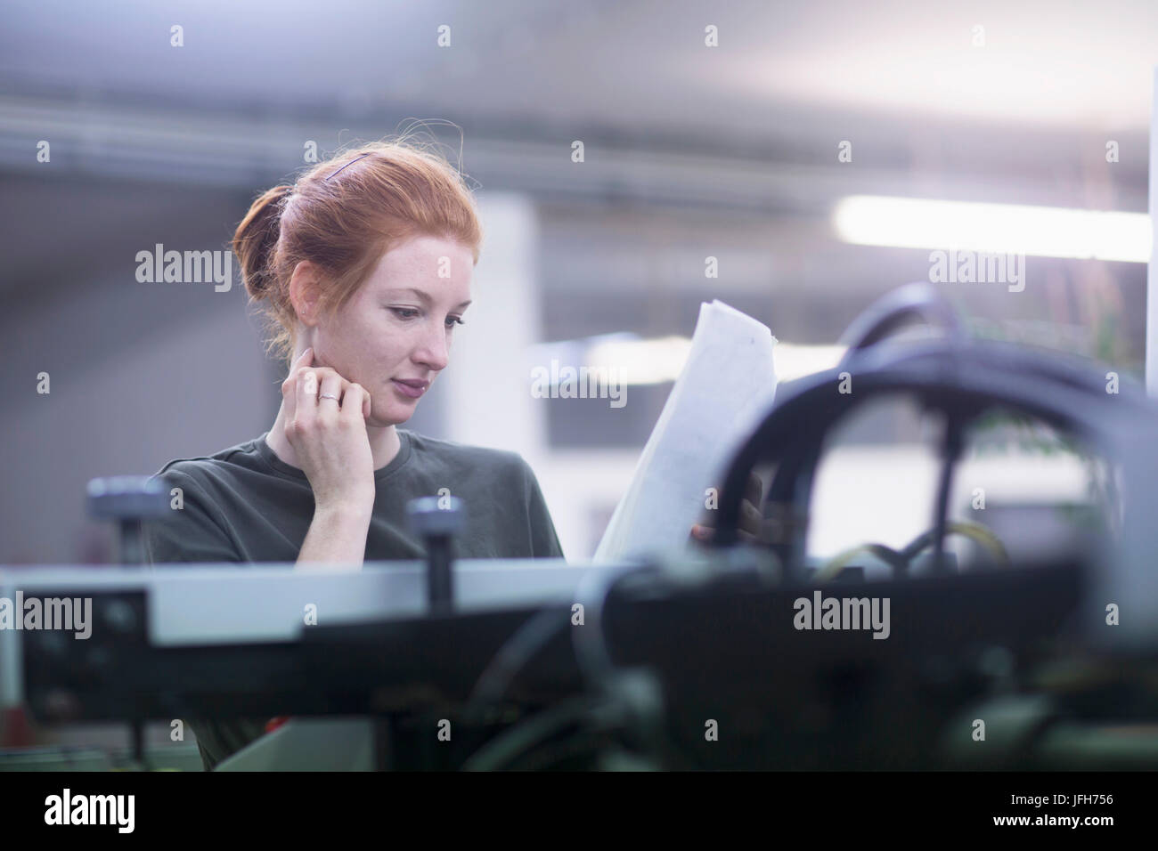 Kontemplative junge Frau liest Papier stehend von Druckmaschinen Stockfoto