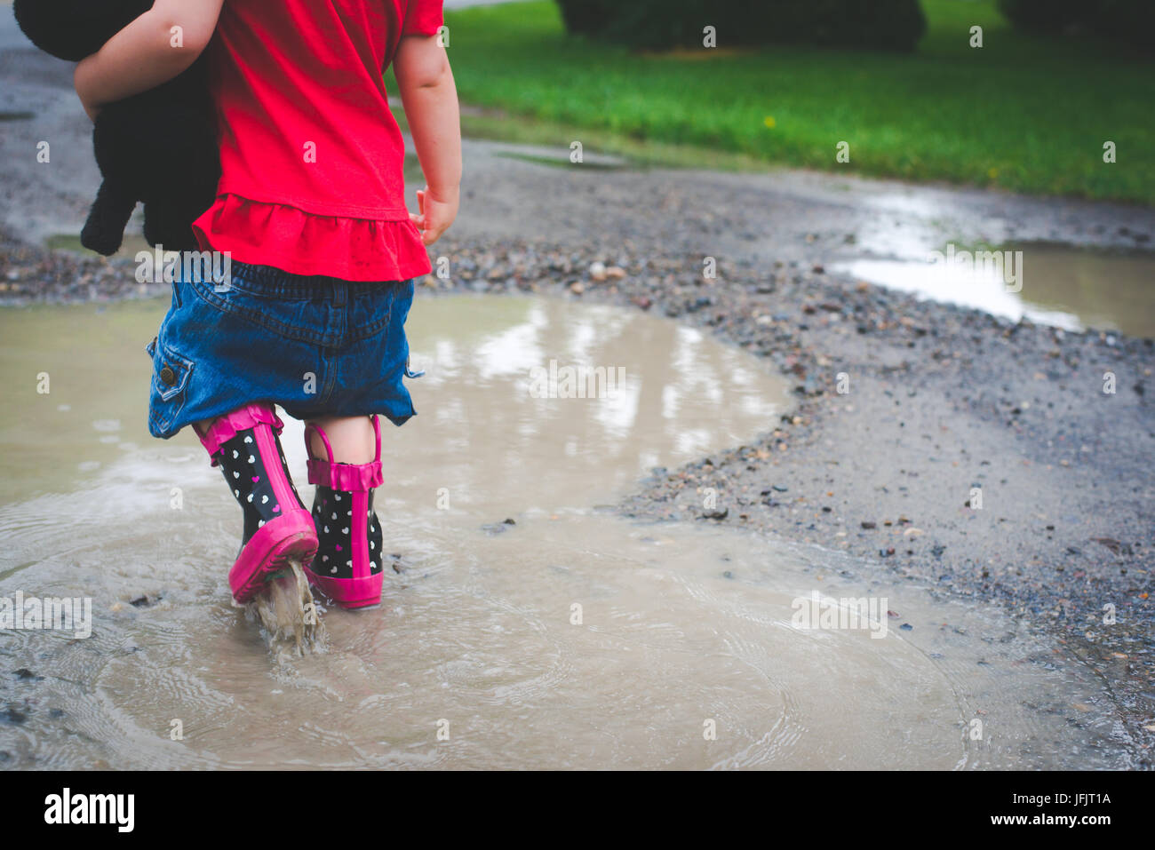 Ein junges Mädchen geht in eine Pfütze mit Regenstiefel auf rot trägt und hält einen kleinen ausgestopften Hund. Stockfoto