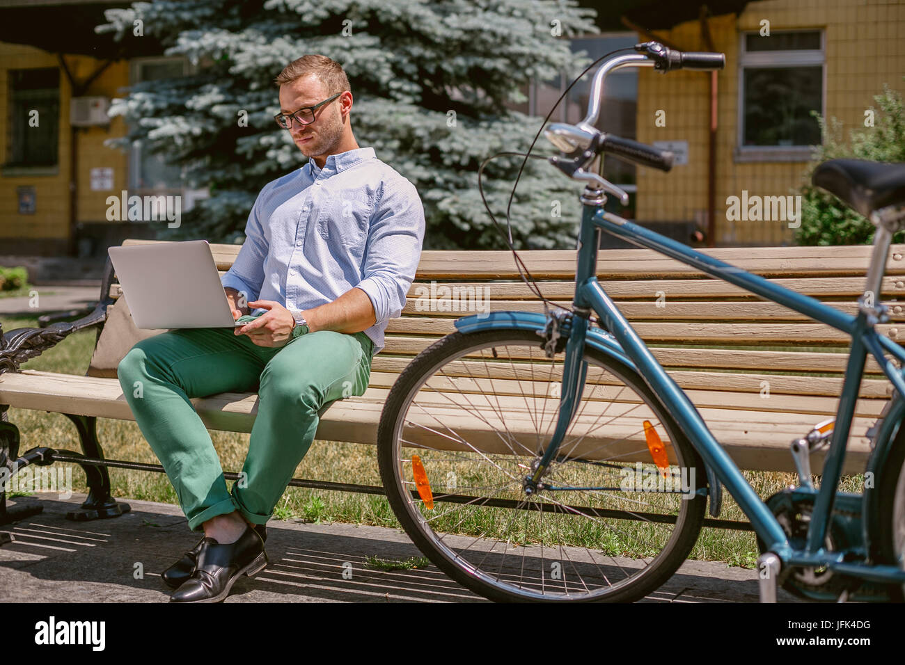 Lässige Geschäftsmann arbeiten mit Laptop auf eine Kaffeepause konzentriert. Er ist auf einer Bank sitzen und arbeiten am Laptop, neben der Bank liegt ein Fahrrad. Stockfoto
