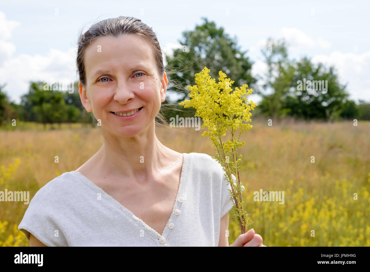 Porträt einer erwachsenen Frau stehend in einer Wiese voller Galium Verum Blumen, auch bekannt als lady's Labkraut oder gelbe Labkraut, mit einem Haufen o Stockfoto