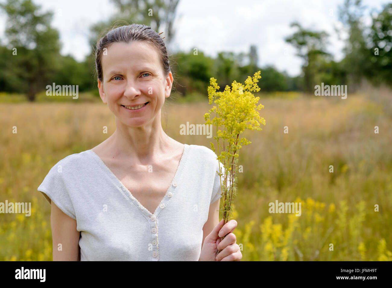 Porträt einer erwachsenen Frau stehend in einer Wiese voller Galium Verum Blumen, auch bekannt als lady's Labkraut oder gelbe Labkraut, mit einem Haufen o Stockfoto