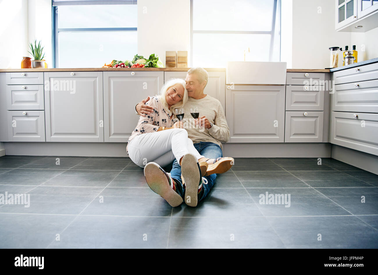 Liebevolle älteres paar lächelnd und zufrieden zusammen auf dem Boden ihrer hellen Küche zu Hause sitzen trinken Rotwein Stockfoto
