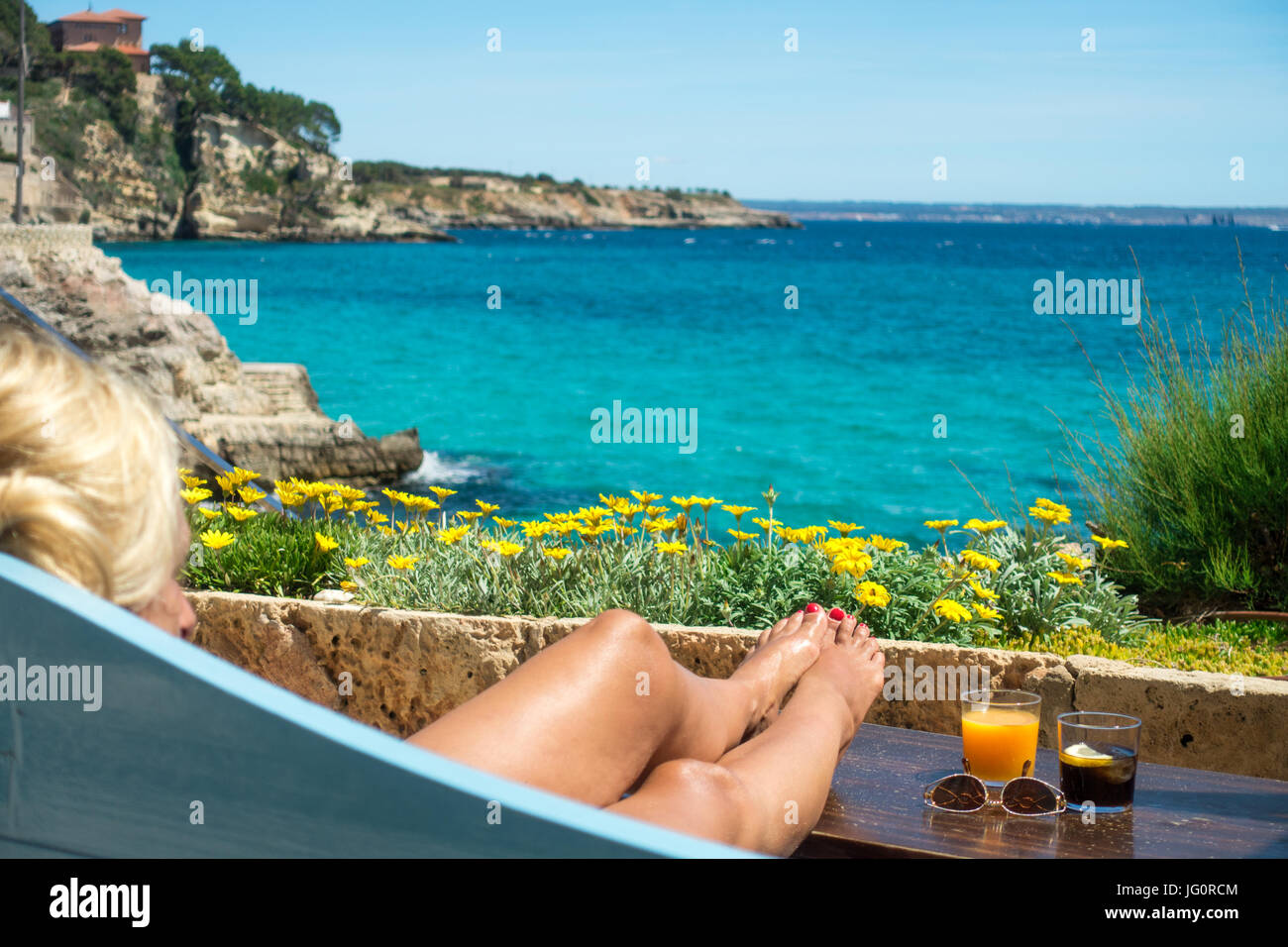 Weibliche Beine und Cocktails vor einem tropischen Meer und Blumen Stockfoto
