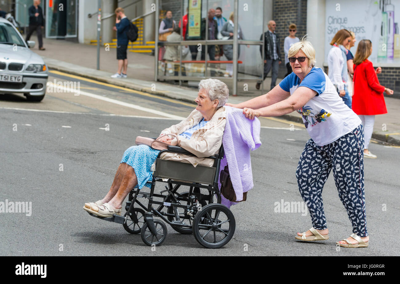 Frau drücken eine ältere Frau in einem Rollstuhl über eine Straße. Stockfoto