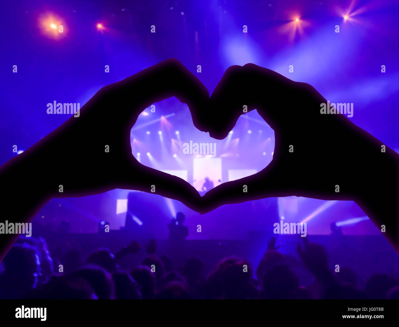 Konzert, Handzeichen in der Form des Herzens für die Musik, verschwommene Publikum und Künstler auf der Bühne im Hintergrund mit blauer Strahler Stockfoto