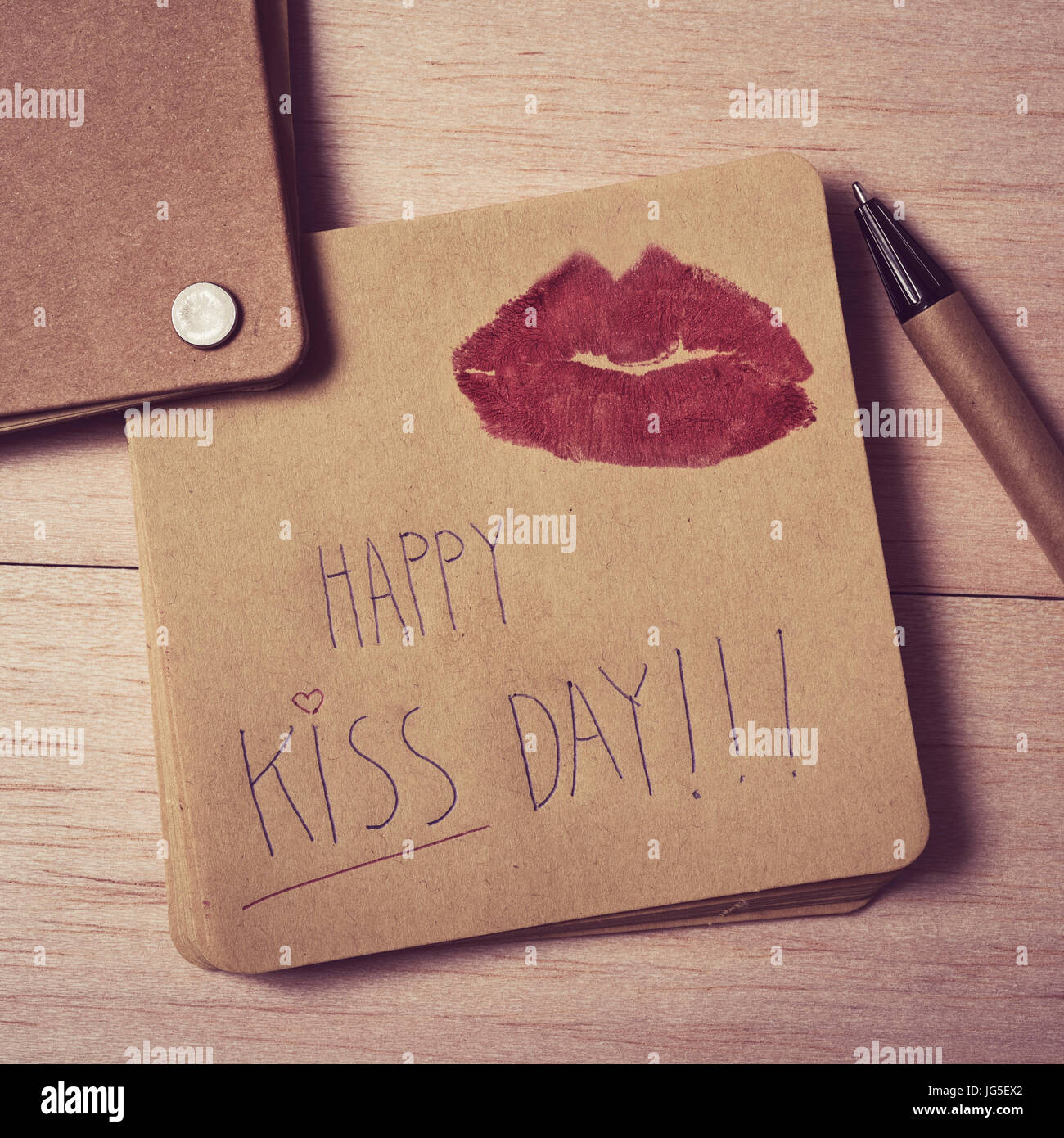High-Angle Shot einer braunen Note mit dem Text zufrieden küssen Tag geschrieben und die Marke von ein paar rote Lippen auf einem Holztisch neben einem Stift Stockfoto