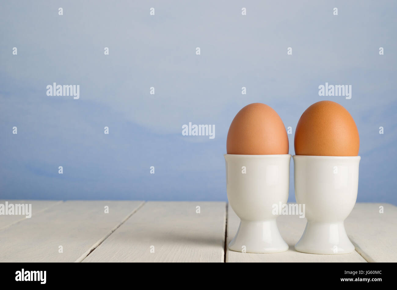Zwei braune undekorierten Frischeier, nebeneinander in weißem Porzellan Eierbecher Hintergrund gemalten blauen Himmel Effekt, auf eine alte Creme lackiert Holz Stockfoto