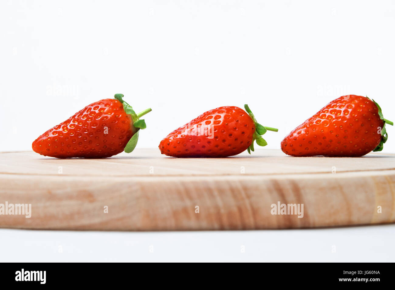Eine Linie von drei roten Erdbeeren mit Blättern und kurz gestielt intakt, sichern Schuss seitlich auf Augenhöhe, auf ein Schneidbrett aus Holz gegen ein cremefarbenes Stockfoto