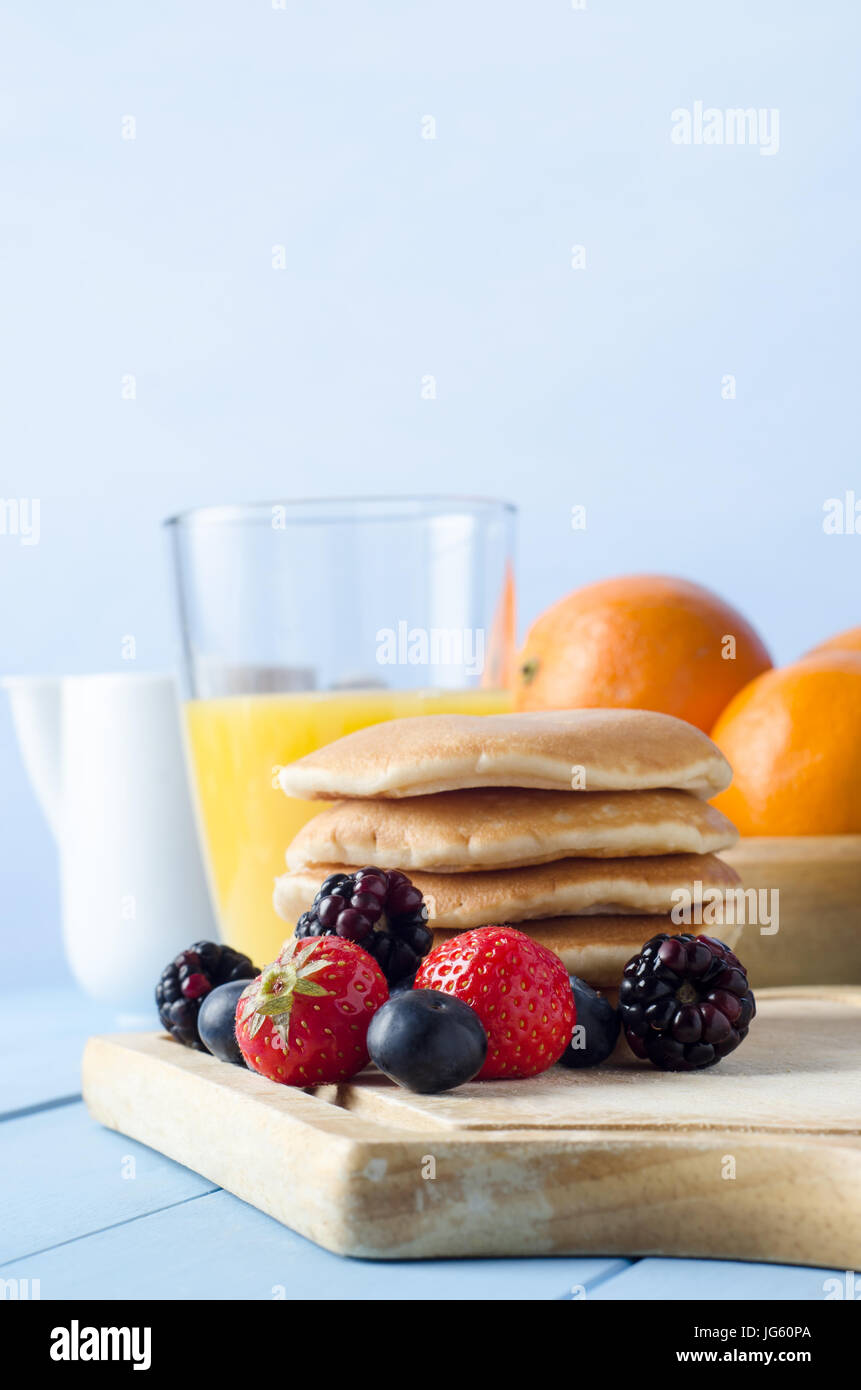 Ein Küchentisch mit Pfannkuchen gestapelt auf Schneidbrett aus Holz umgeben von Sommerfrüchten, ein Glas Orangensaft, Schale von Orangen und Milch Stockfoto