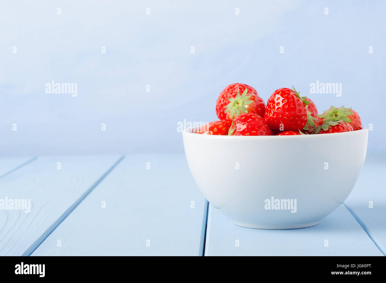 Eine enge, Auge Ebene Seitenansicht einer Weißware Schüssel Erdbeeren auf der rechten Seite des Rahmens, auf einem leichten blau lackierten Holzbohle Tisch mit blauen backgr Stockfoto