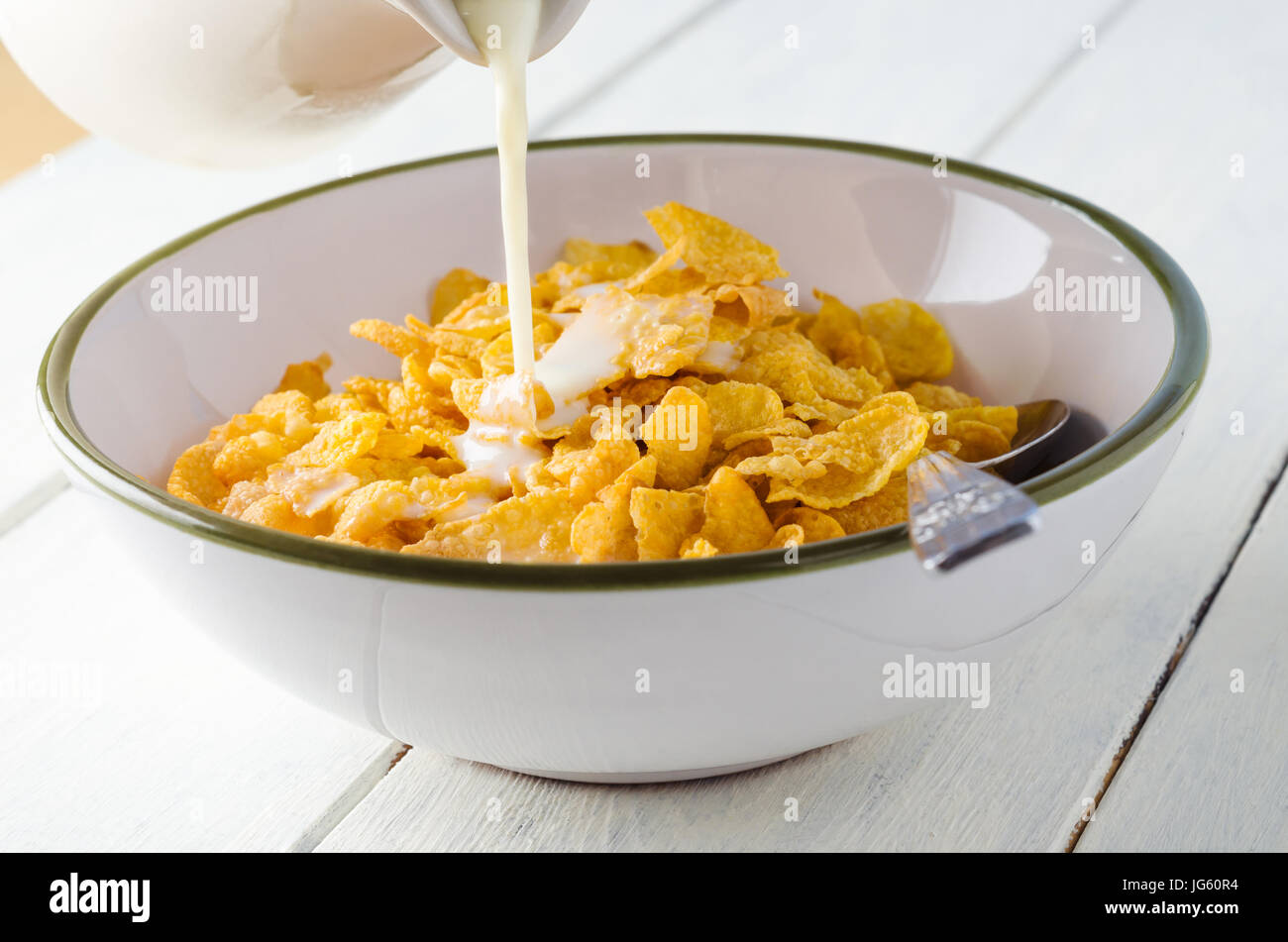 Milch gießen aus einem Krug in eine Schüssel mit Frühstück Cornflakes, auf einem weiß lackierten Holz beplankt Küchentisch. Stockfoto