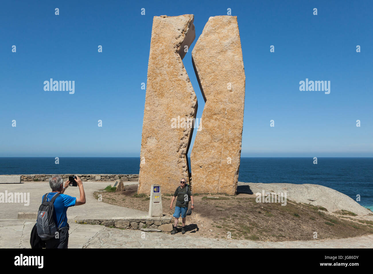 Menschen fotografieren vor der Skulptur Hommage "Pedra da Ferida" ("Wunde Stein") von spanischen Bildhauers Alberto Bañuelos gewidmet die Freiwilligen, die geholfen haben, reinigen Sie die Prestige-Ölpest im Jahr 2002 an der Küste des Atlantischen Ozeans, bekannt als der Costa De La Muerte (Todesküste), in der Nähe der Stadt Muxía in Galicien, Spanien. Der Meilenstein des Camino de Santiago (Jakobsweg) im Vordergrund zu sehen. Stockfoto