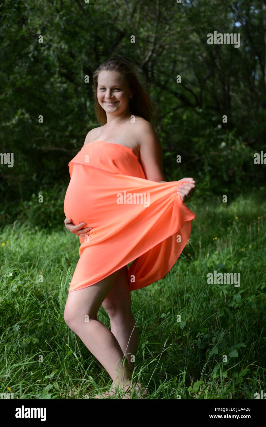 Schwangere junge Frau mit orange Kleid steht in einem park Stockfoto