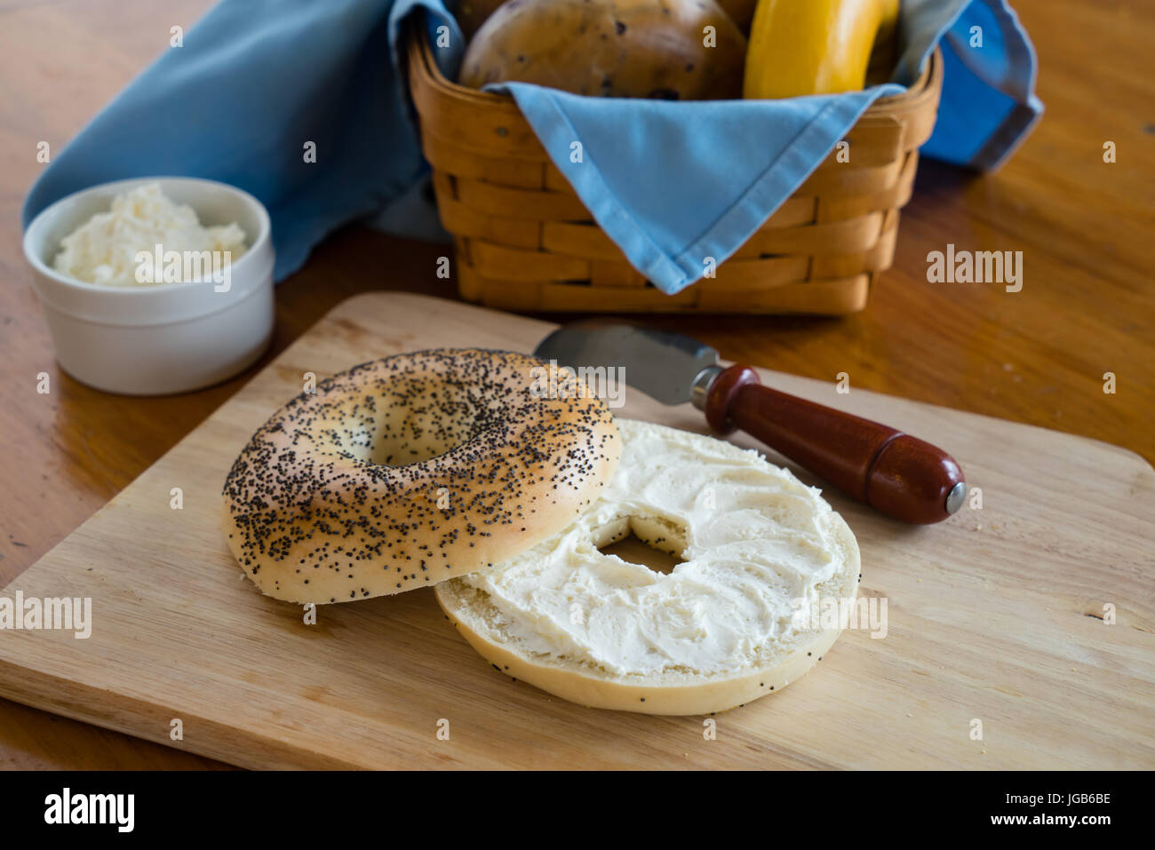 Mohn-Bagel mit Frischkäse mit einem Korb von sortierten Bagels im Hintergrund. Stockfoto