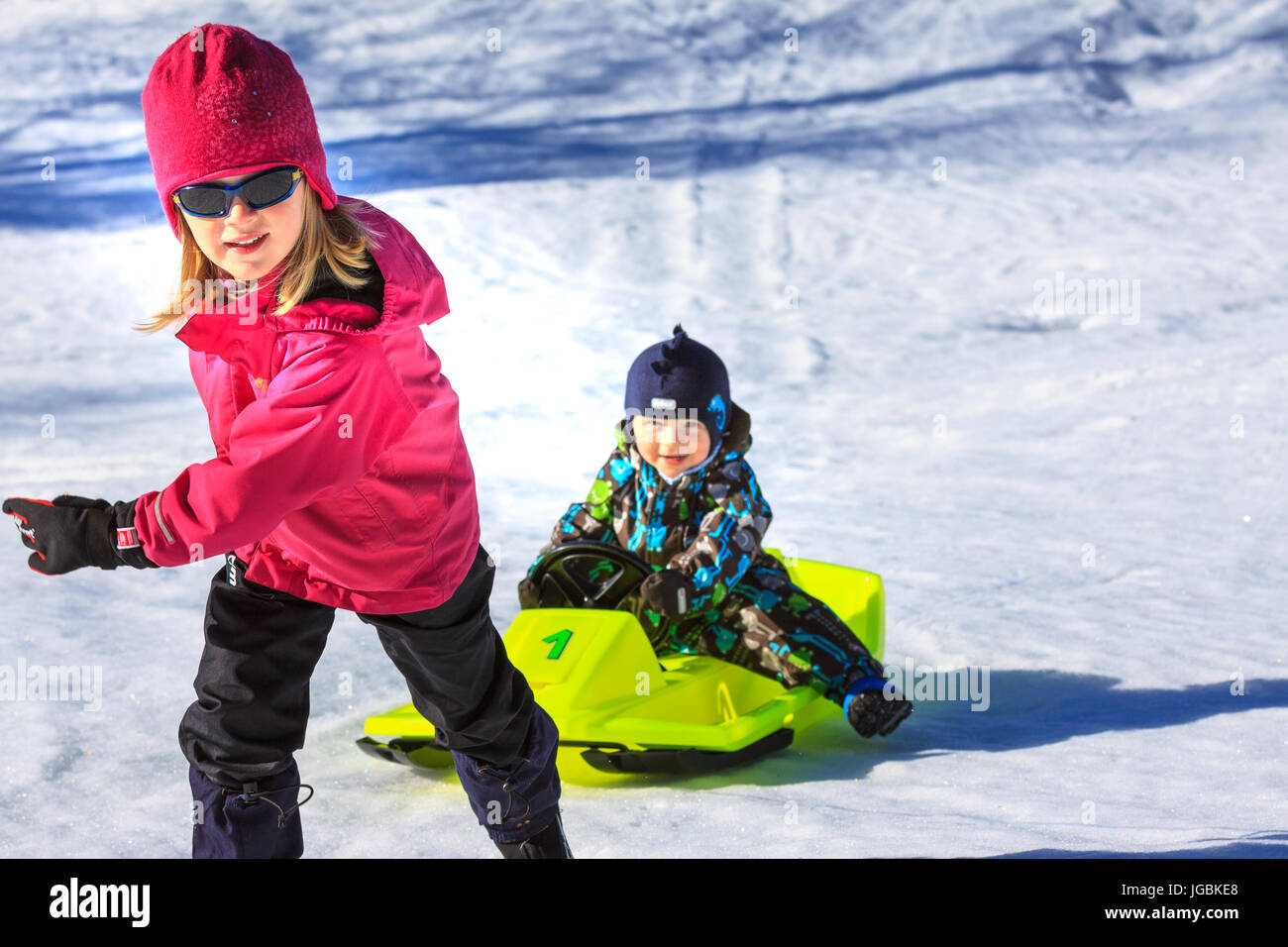 Mädchen ziehen jüngeres Kind auf gelben Rodel Schlitten, verschneite Hügel hinauf. Knackige Farben. Stockfoto