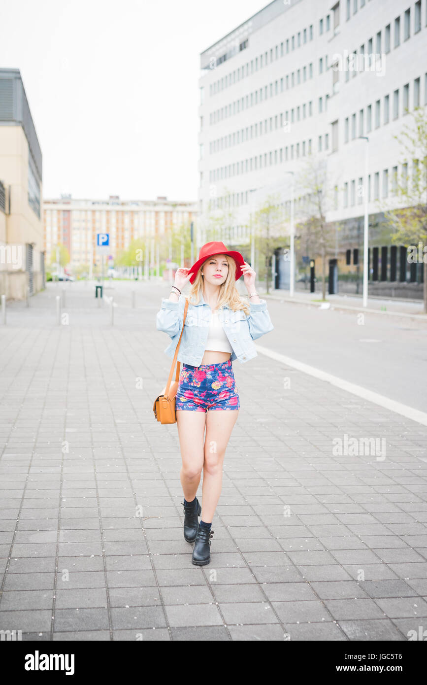 Junges schönes blonde kaukasisches Mädchen zu Fuß in die Vororte der Stadt bekleidet mit einem roten Hut, ein paar Blumen Shorts und Jeansjacke Stockfoto