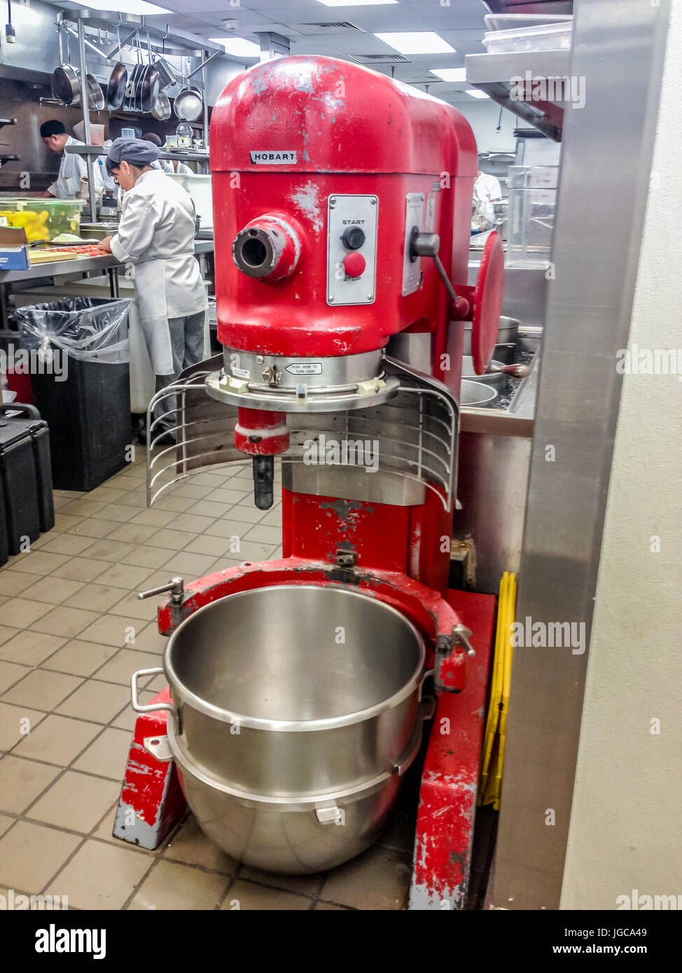 Große rote industrielle Mischer mit einer leeren sauberen Edelstahlschüssel in einer Bäckerei mit Menschen arbeiten bei Küchenarbeitsplatten im Hintergrund Stockfoto