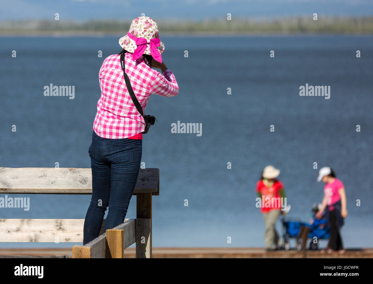 Eine Frau trägt ein rosa karierte Bluse und einen Hut mit einer rosa Schleife nimmt Fotos von ihrer Familie in West Thumb Geyser Basin im Yellowstone National Park Stockfoto